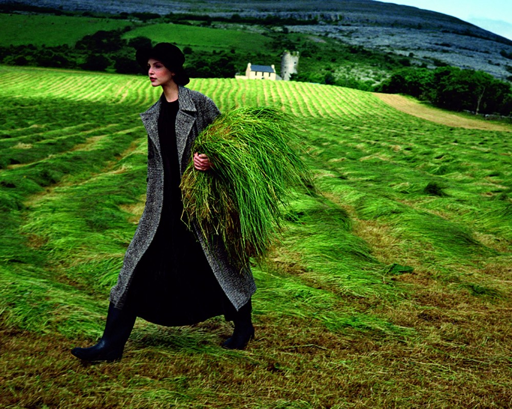 Model Nadja Auermann wearing a long tweed coat walking through a field carrying long grass VOGUE September 1993.